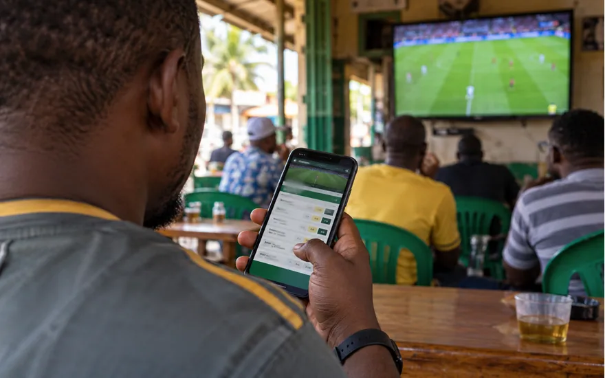 adult man from behind holding a phone with blurred sports bet slip in a Dar es Salaam sports bar; distant football television; your first betPawa session. Illustrative generated scene.