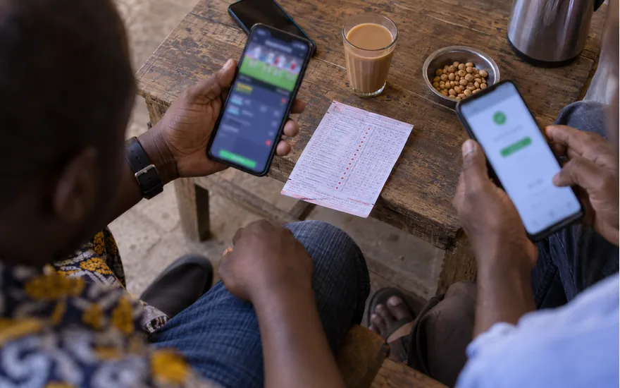 overhead of adult Tanzanian hands with two phones, one blurred sports slip and one blurred mobile money confirmation beside a paper betting ticket; your first betPawa session. Illustrative generated scene.